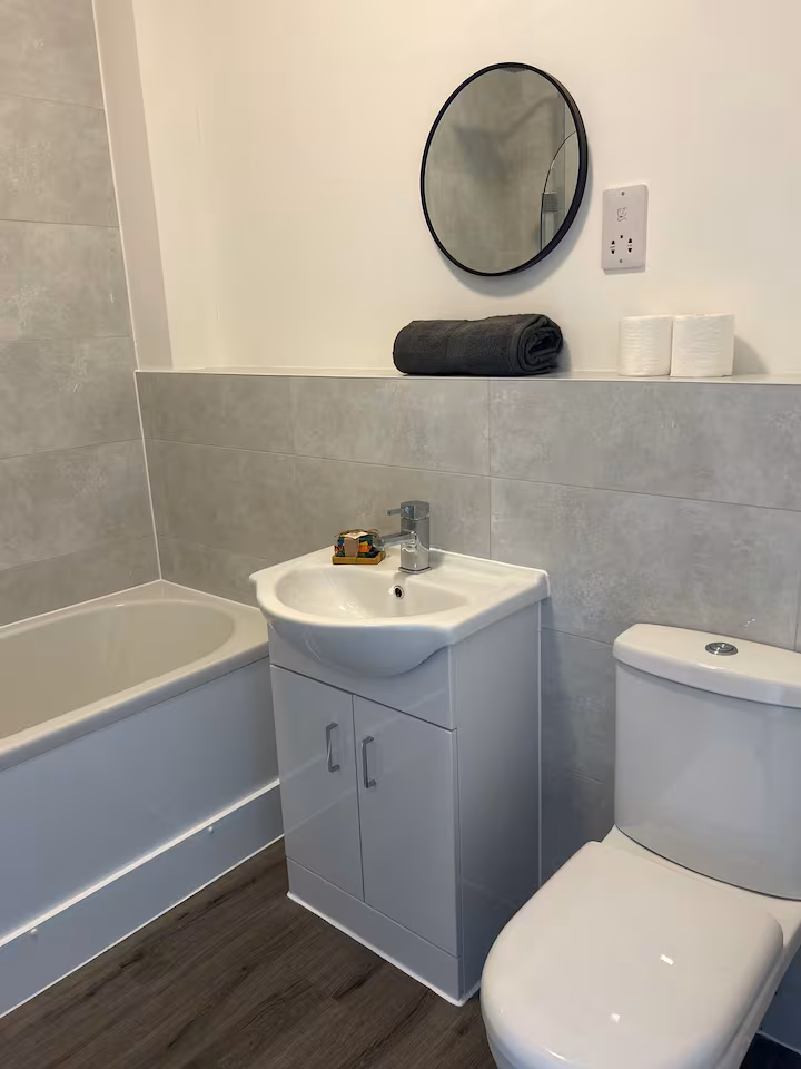 Bright, shiny, inviting bathroom with toilet, white sink and cupboard, bathtub, grey wall tiles, and laminate flooring