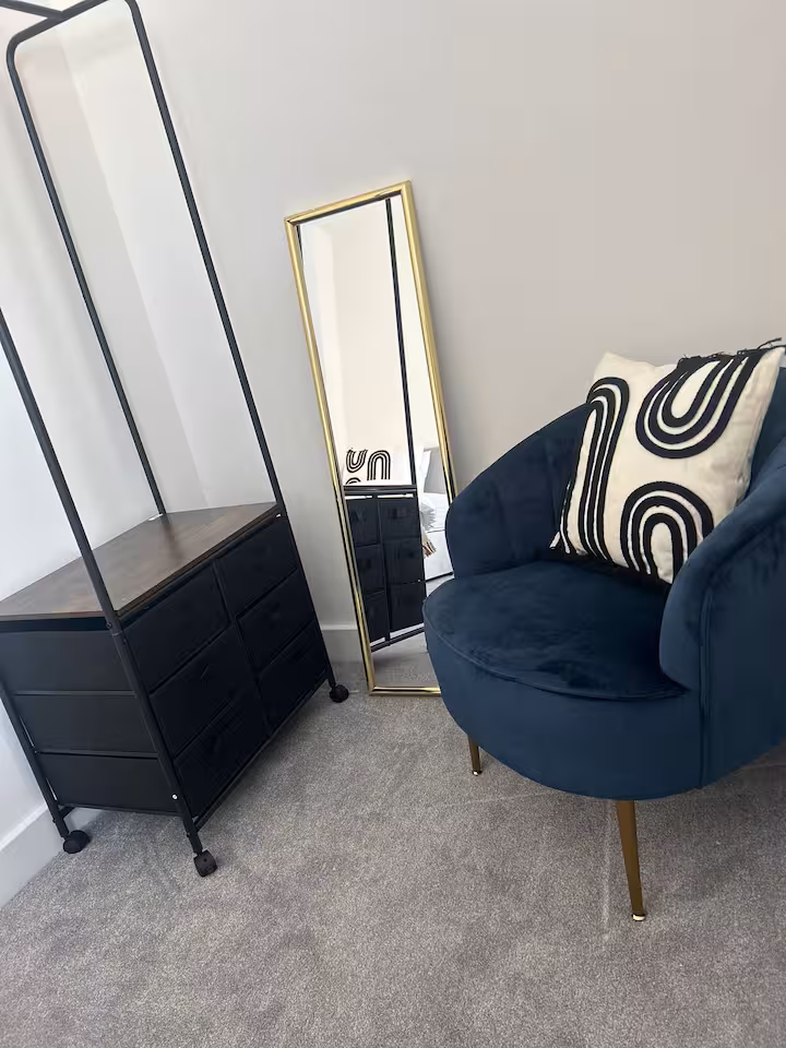Black clothes rail with drawers, rectangular mirror, and comfy luxurious blue chair with cushion located in a bright, airy bedroom