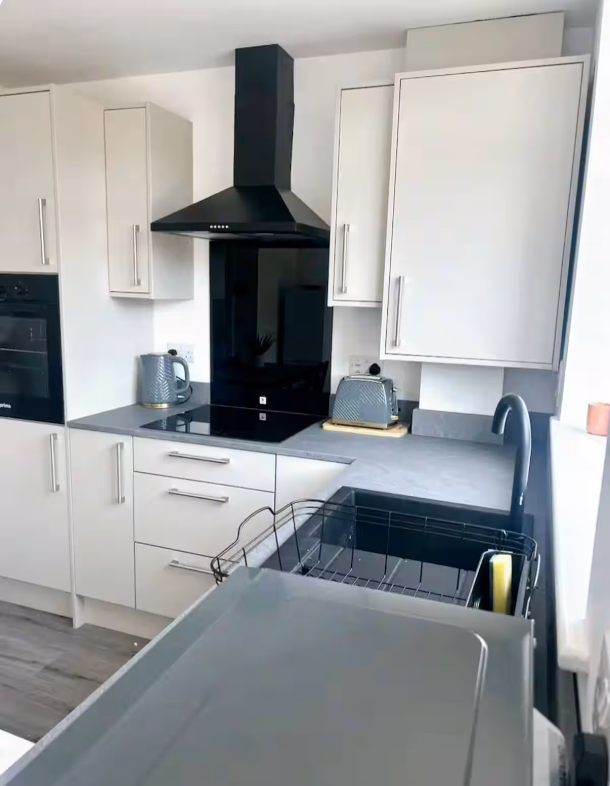 Bright, modern kitchen featuring white cupboards, black oven, extractor fan, electric stove, grey kettle and toaster, sink, and laminate flooring
