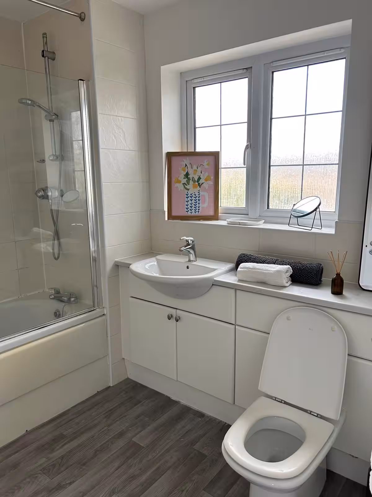 Bright, airy bathroom with bathtub and shower, sink with cupboard, toilet, laminate flooring, and pretty decorations