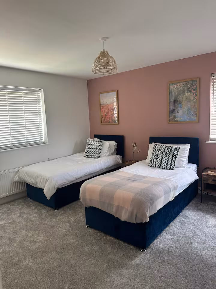 Two single beds located in a cozy bedroom with soft, fluffy carpet, white blinds, and pleasant wall art
