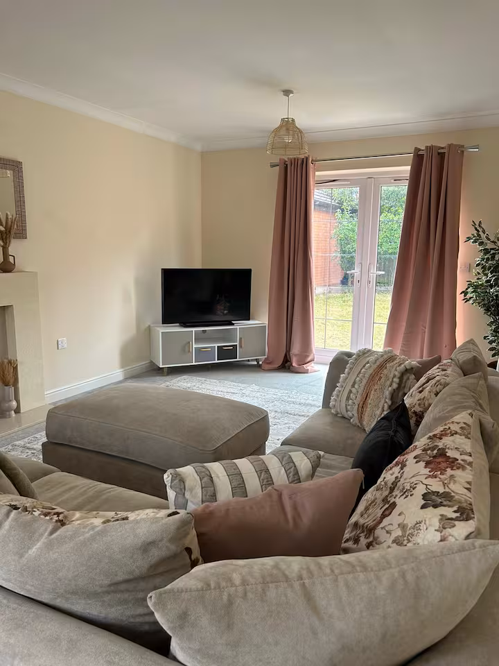 Spacious living room with large comfy sofa and cushions, square foot rest, standing television, and pink curtains