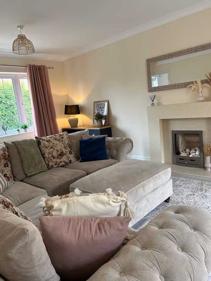 Large traditional sofa, plentiful of cushions, located in a spacious living room with foot rest, table lamp, and silver fireplace